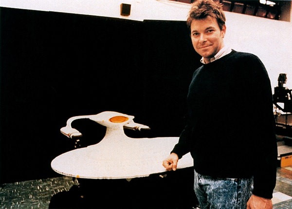 A very young-looking Johnathan Frakes ("Will Riker") poses beside an upside-down model of the Enterprise-D