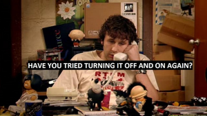 Still photo from “The IT Crowd” — Rory at his desk on the phone, with the caption “Have you tried turning it off and on again?”