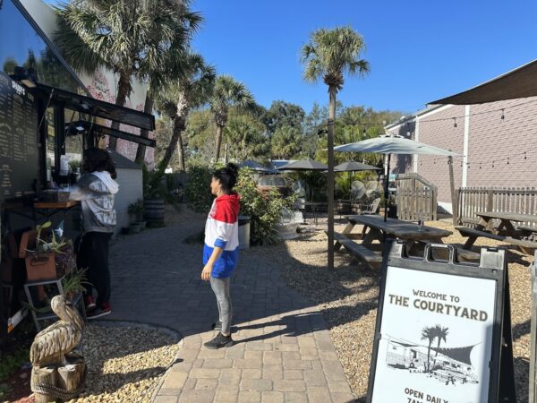 The courtyard at Spaddy’s Coffee in Seminole Heights, Tampa. It’s an open space sandwiched between two one-storey buildings with picnic and patio tables, and palm trees. The sky is blue and cloudless. There is a line for coffee at the trailer.