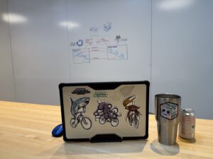 Joey’s laptop and insulated mug sitting on a table, with Embarc Collective’s tall whiteboards in the background.